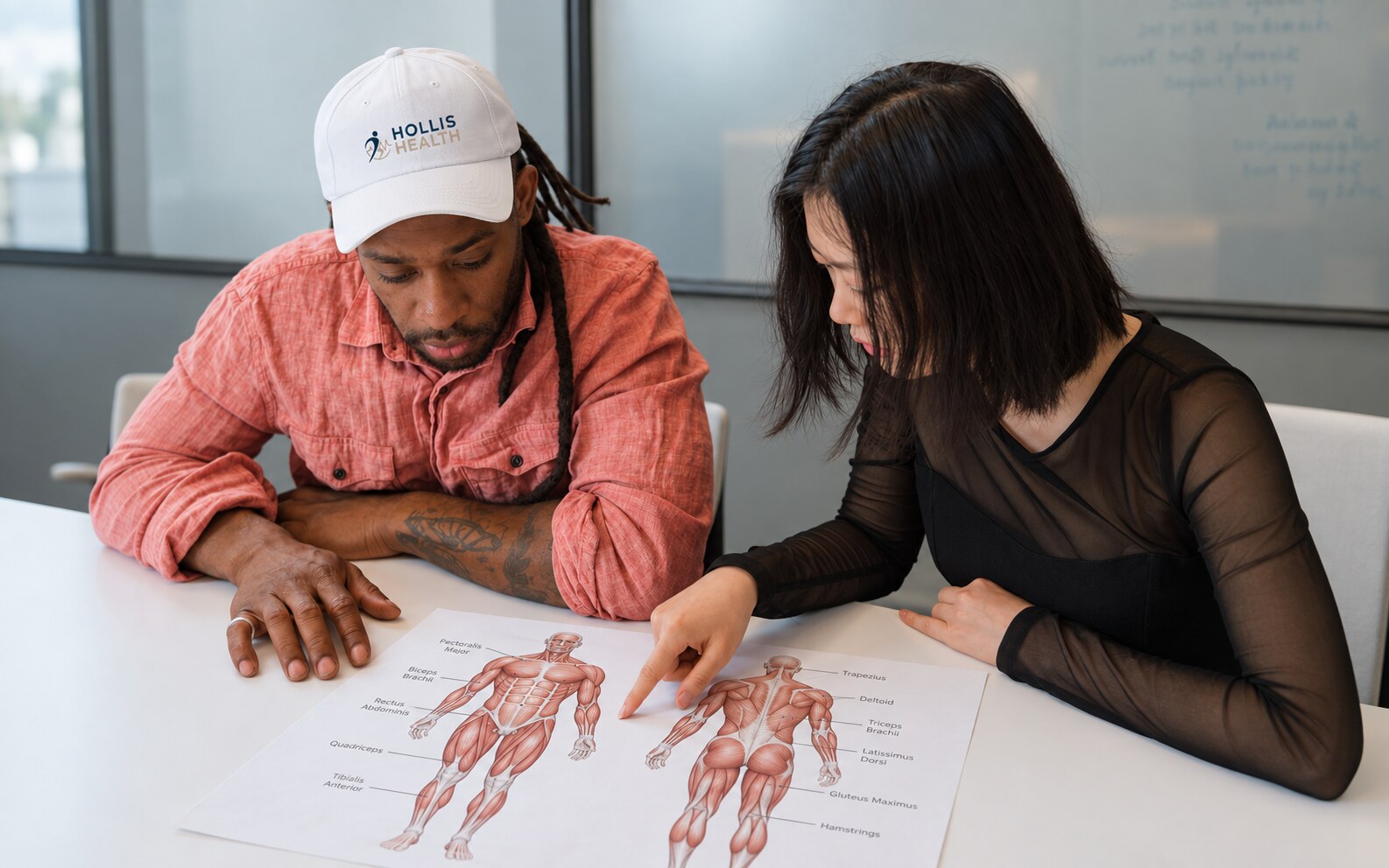 Two people reviewing a muscle anatomy chart, one wearing a Hollis Health cap